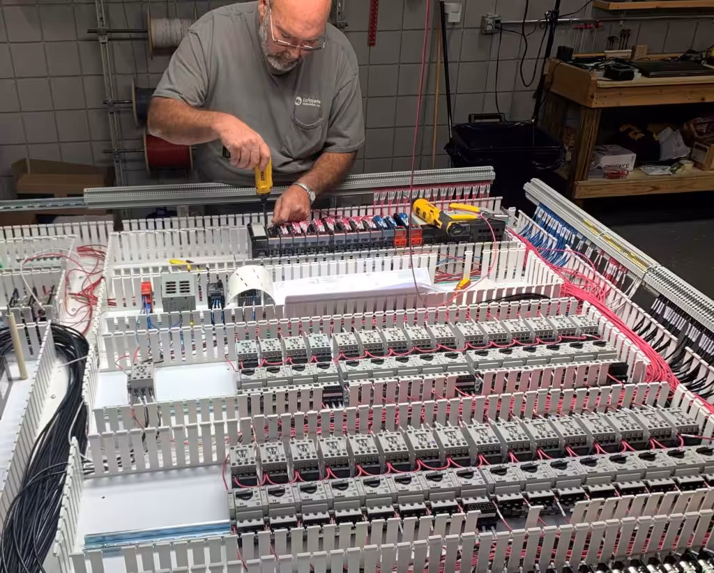 Technician wiring a large control panel lined with rows of relays, contactors, and terminal blocks